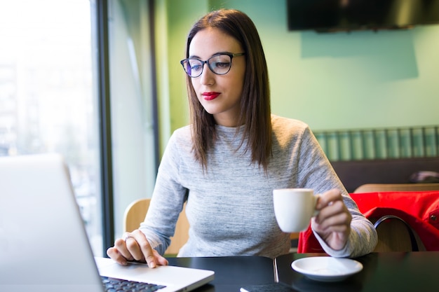 Caffè bevente della bella giovane donna mentre per mezzo del suo computer portatile nella caffetteria.