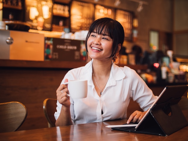 Caffè bevente della donna asiatica in caffè e computer portatile usando per l&#39;affare lavorante