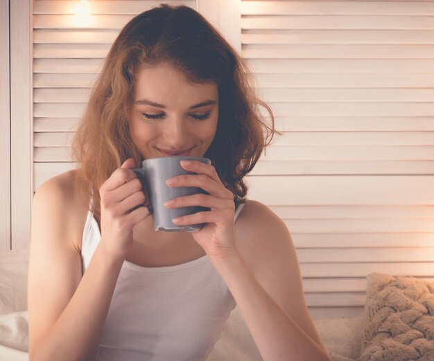 Caffè bevente della donna graziosa che si siede sul letto a casa