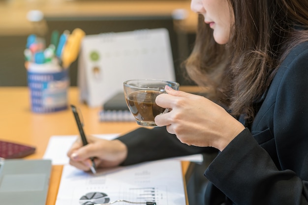 Caffè bevente della mano della donna di affari del primo piano quando lavorano con il computer portatile del computer e il supplie dell&#39;ufficio