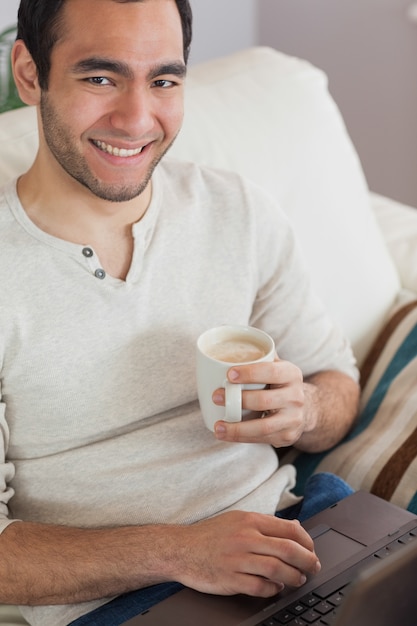 Caffè bevente sorridente dell&#39;uomo attraente mentre usando il suo computer portatile