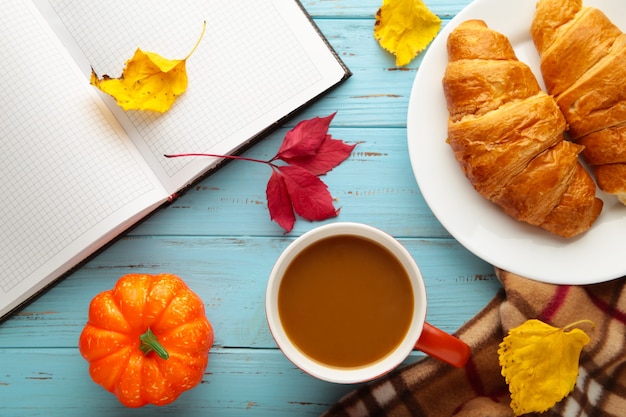 Caffè caldo con croissant e foglie autunnali su blu - concetto di relax stagionale. Vista dall'alto