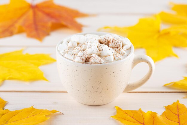 Caffè caldo con marshmallow in una tazza bianca su un tavolo di legno con foglie autunnali
