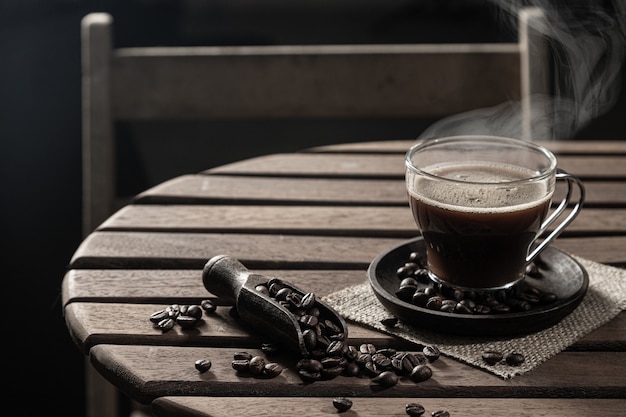 Caffè caldo in una tazza di vetro e chicchi di caffè su un tavolo di legno su sfondo scuro.