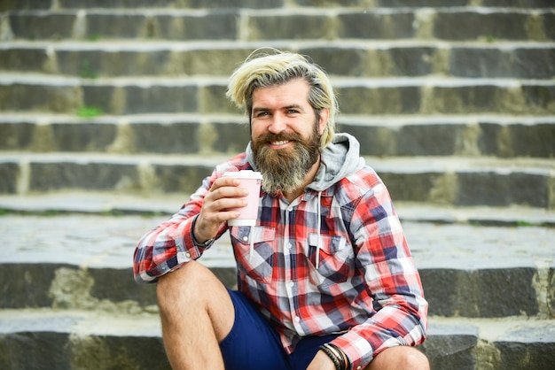 Caffè da asporto Puro piacere Dose di caffeina Buon umore Cultura del caffè della terza ondata Uomo con la barba che beve caffè Uomo barbuto che beve caffè sfondo urbano Hipster rilassarsi sulle scale Riposarsi