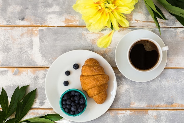 Caffè del mattino, cornetto, mirtilli e bellissimi fiori gialli di peonia sul tavolo luminoso