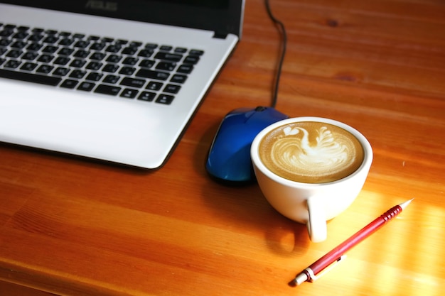 Caffè del mattino sul tavolo dell'operaio. luce e ombra sulla tazza da caffè bianca sul pavimento in legno con sfocatura dello sfondo del laptop