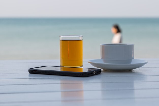 Caffè del tè con lo Smart Phone sulla tavola di legno bianca ai precedenti della spiaggia