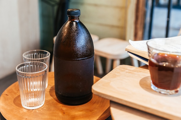 Caffè espresso freddo dentro la bottiglia nera sulla tavola di legno con i bicchieri vuoti.