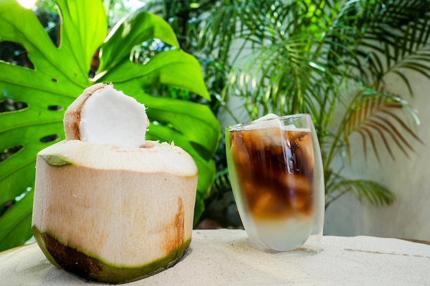 Caffè ghiacciato al cocco in vetro a doppia parete sulla sabbia su sfondo verde foglia