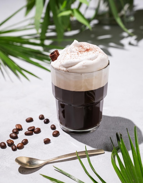 Caffe' ghiacciato in un bicchiere con schiuma su uno sfondo chiaro con foglie di palma, chicchi di caffè e ombre mattutine