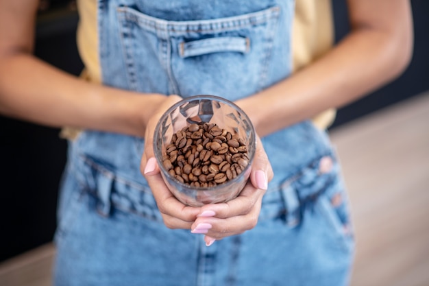 Caffè in grani interi. Mani femminili graziose con manicure che tiene un bicchiere di chicchi di caffè belli, senza volto