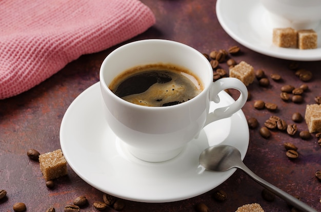 Caffè in tazza e piattino su sfondo scuro con chicchi di caffè e zucchero di canna.