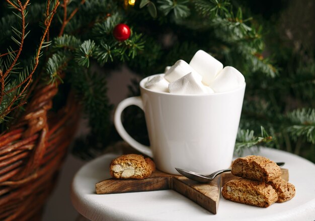 Caffè in una tazza bianca con marshmallow. Caffè festivo mattutino con i tradizionali cantuccini italiani alle mandorle. Una tazza di caffè su uno sfondo di rami di abete verde su un supporto bianco.