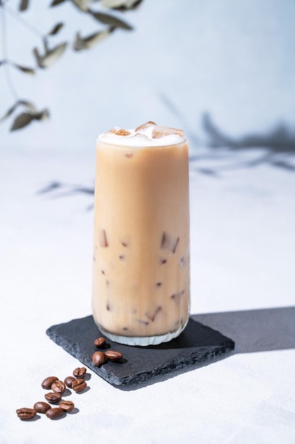 Caffè latte ghiacciato in un bicchiere alto con latte su uno sfondo chiaro con chicchi di caffè e ombre mattutine Concetto di rinfresco estivo