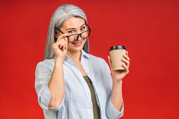 Caffè mattutino per andare Affascinante donna anziana in abbigliamento formale in piedi con tazza di carta di bevanda calda isolata su sfondo rosso che si prepara a una buona giornata di lavoro Donna d'affari matura prende una pausa caffè