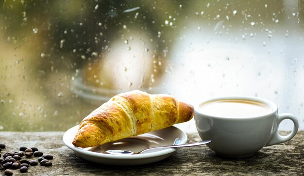 Caffè preparato fresco in tazza bianca o tazza sul davanzale. Finestra di vetro bagnata e tazza di bevanda calda alla caffeina. Bevanda al caffè con dolce croissant. Godendo il caffè in una giornata di pioggia. L'ora del caffè in una giornata di pioggia.