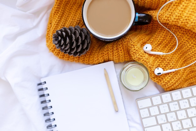 Caffè, taccuino pulito. tastiera, cono, candela, auricolari su fogli bianchi stropicciati e vista dall'alto del coperchio a maglia gialla. Donna che lavora a casa. Colazione accogliente. Modello. Stile piatto.