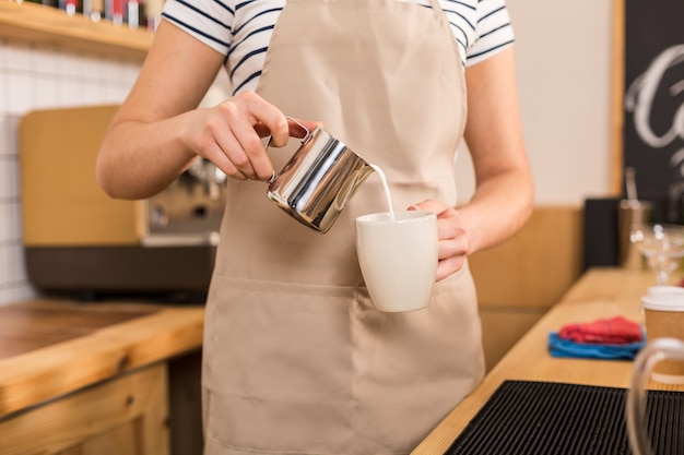 Caffèlatte. Primo piano di latte che viene versato nella tazza da una donna abile felice piacevole mentre si lavora nella caffetteria