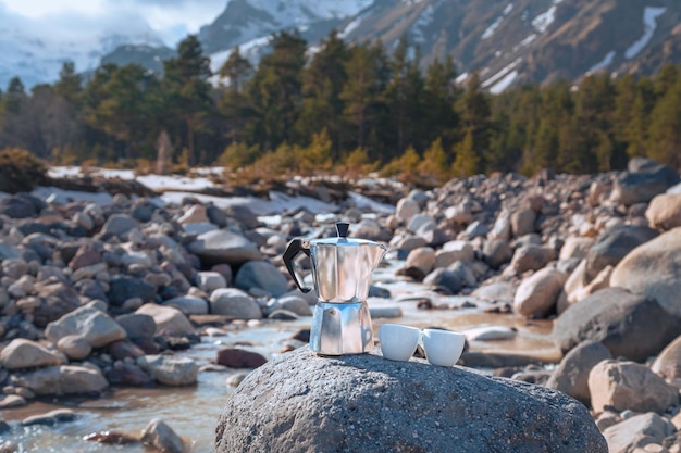 Caffettiera Moka e due tazze in porcellana su pietra grande sullo sfondo del fiume di montagna