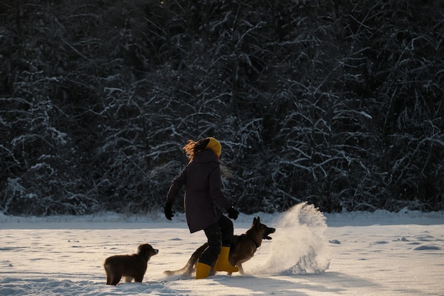 Calciare la neve con i piedi Donna felice in passeggiata nel parco invernale che gioca con i cani