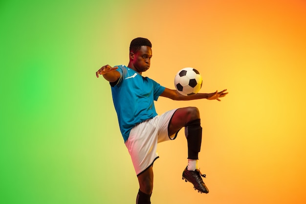 Calcio maschio afroamericano, allenamento del giocatore di football in azione isolato su sfondo sfumato studio in luce al neon. Concetto di movimento, azione, risultati, stile di vita sano. Cultura giovanile.
