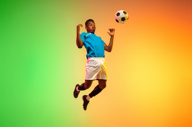 Calcio maschio afroamericano, allenamento del giocatore di football in azione isolato su sfondo sfumato studio in luce al neon. Concetto di movimento, azione, risultati, stile di vita sano. Cultura giovanile.