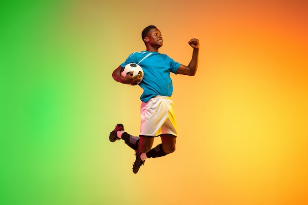 Calcio maschio afroamericano, allenamento del giocatore di football in azione isolato su sfondo sfumato studio in luce al neon. Concetto di movimento, azione, risultati, stile di vita sano. Cultura giovanile.