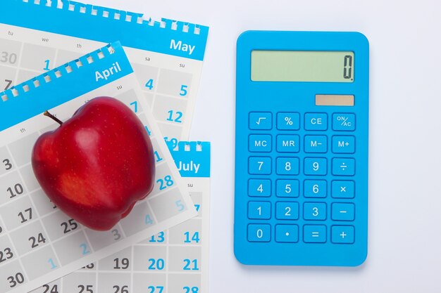 Calcolatrice con i fogli del calendario mensile, mela rossa su fondo bianco. Calcolo economico, costi