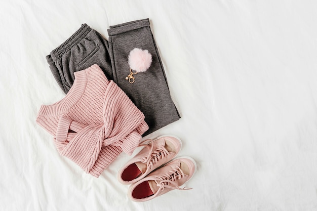 Caldo maglione rosa pallido e pantaloni grigi con scarpe da ginnastica su lenzuolo bianco. Elegante outfit da donna autunnale o invernale. Collage di vestiti alla moda. Disposizione piana, vista dall'alto.