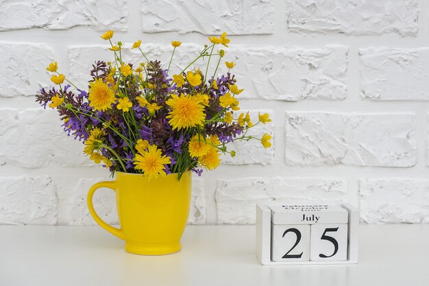 calendario 25 luglio e tazza gialla con fiori colorati luminosi contro il muro di mattoni bianco.