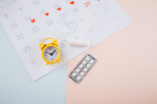 Calendario delle mestruazioni con allarme giallo, tampone di cotone e pillole contraccettive su sfondo rosa. Giorni critici della donna, concetto di protezione dell'igiene della donna.