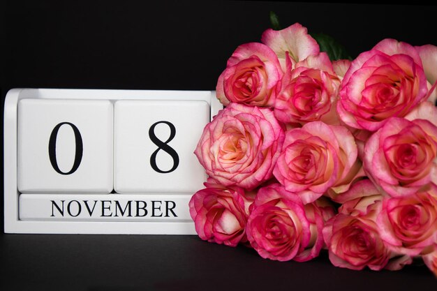 Calendario in legno dell'8 novembre, bianco su sfondo nero, rose rosa nelle vicinanze