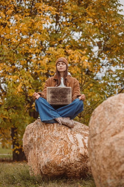 Calma freelance femminile con laptop che medita sulla roccia in natura in autunno durante il lavoro a distanza