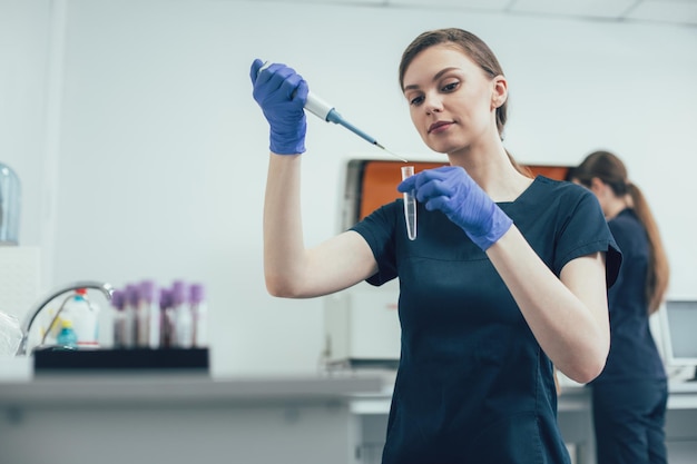 Calma giovane donna con guanti di gomma che tiene in mano una provetta sterile e vi fa cadere una sostanza con un erogatore di pipette