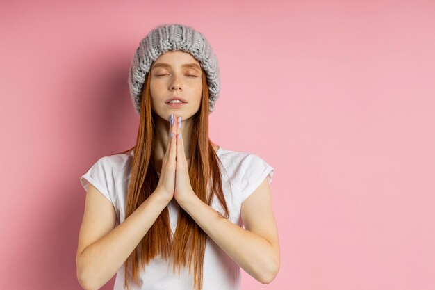 Calma giovane donna dai capelli rossi con gli occhi chiusi che pratica yoga, tiene i palmi uniti, prega per la pace e l'amore, medita, indossa un cappello lavorato a maglia grigio e una maglietta bianca su sfondo rosa con spazio per le copie