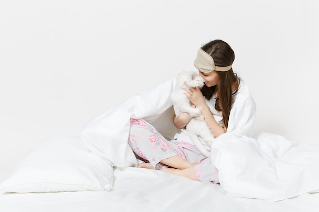 Calma giovane donna seduta a letto con maschera per dormire sugli occhi isolati su muro bianco