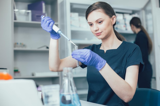 Calma giovane donna seduta al suo posto di lavoro in laboratorio e tenendo in mano una provetta mentre misura il liquido con un erogatore automatico di pipette