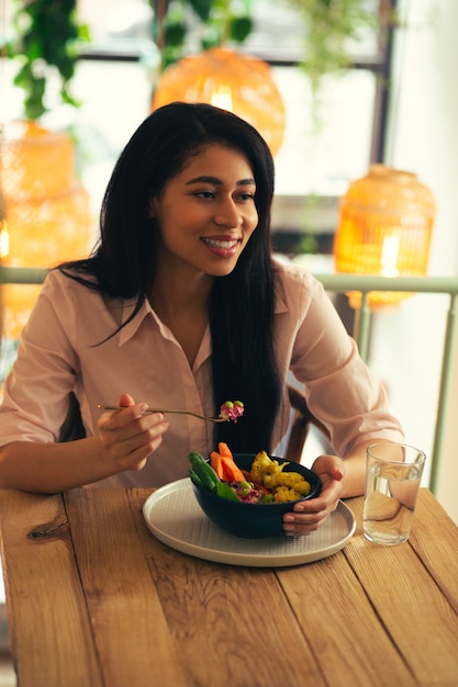 Calma giovane donna seduta al tavolo di legno in un bar e guardando lontano con un sorriso mentre mangia il suo pasto vegetariano