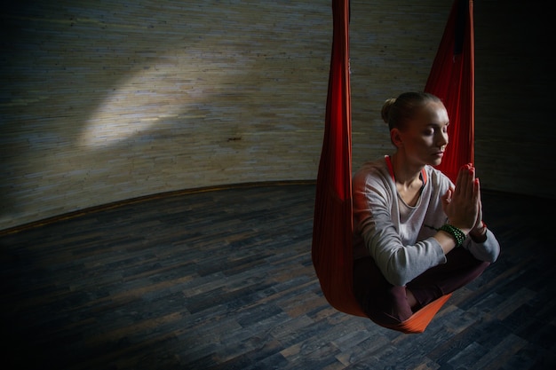Calma giovane donna seduta in amaca arancione con le mani davanti al petto e chiudendo gli occhi mentre praticava lo yoga aereo