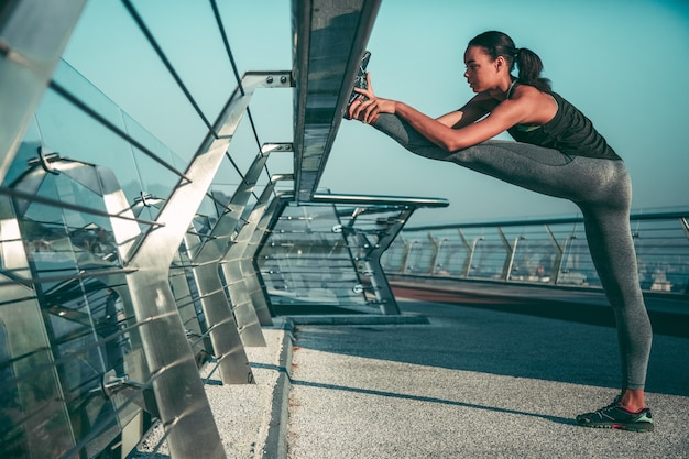 Calma giovane sportiva che mette piede sulla ringhiera del ponte e scalda i muscoli