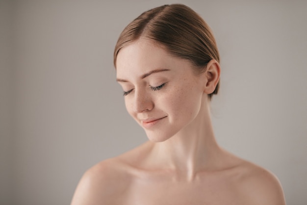 Calmo e bello. Ritratto di donna dai capelli rossi con le lentiggini che tiene gli occhi chiusi e sorride mentre sta in piedi su uno sfondo grigio