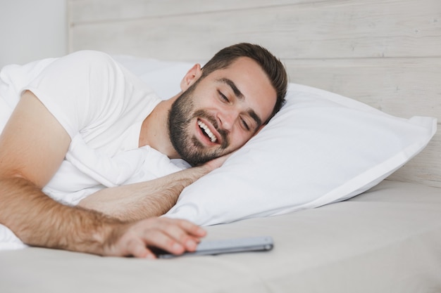 Calmo giovane bell'uomo barbuto sdraiato a letto con una coperta di cuscino in lenzuolo bianco in camera da letto a casa