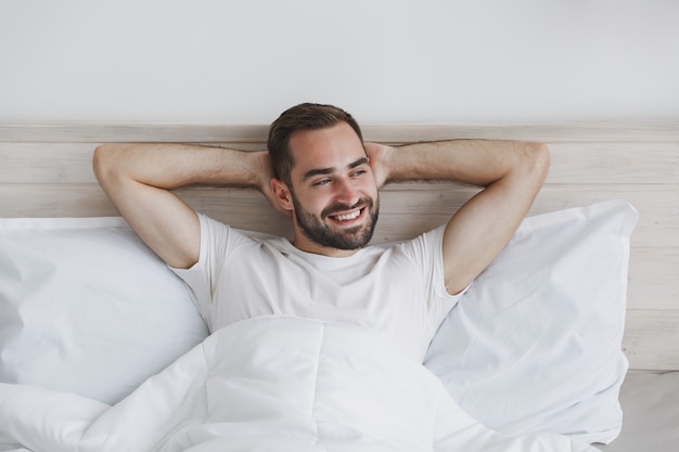 Calmo giovane bell'uomo barbuto sdraiato a letto con una coperta di cuscino in lenzuolo bianco in camera da letto a casa