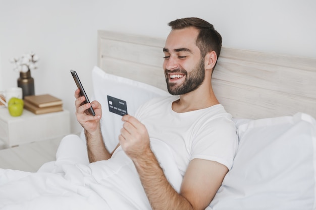 Calmo giovane uomo barbuto sdraiato a letto con coperta di cuscino lenzuolo bianco in camera da letto a casa