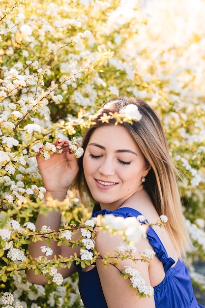 Calmo sensuale ritratto di donna bella hipster in cespuglio fiorito con fiori bianchi di spirea. Copia spazio. Elegante donna bionda in abito blu in giardino