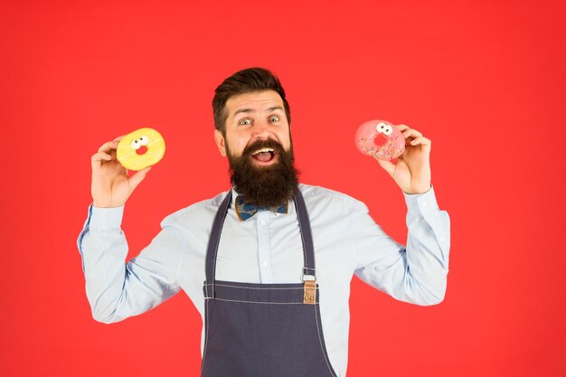 Calorie della ciambella Ciambella smaltata Uomo barbuto ben curato in grembiule che vende ciambelle Cibo per ciambelle Prodotti da forno Dolci e torte Cibo spazzatura Hipster panettiere barbuto tenere ciambelle Ciambella dolce Cameriere nella caffetteria