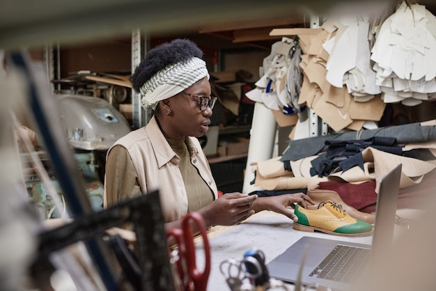 Calzolaio africano seduto al suo posto di lavoro con modelli e facendo nuove scarpe in officina