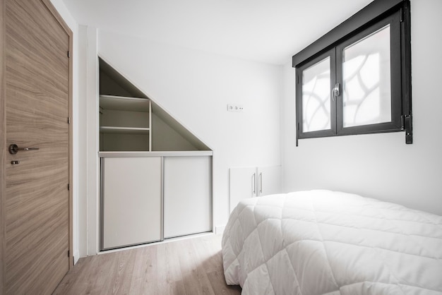 Camera con letto ad angolo coperto da piumino bianco trapuntato alluminio nero finestra porta legno e armadio a forma triangolare