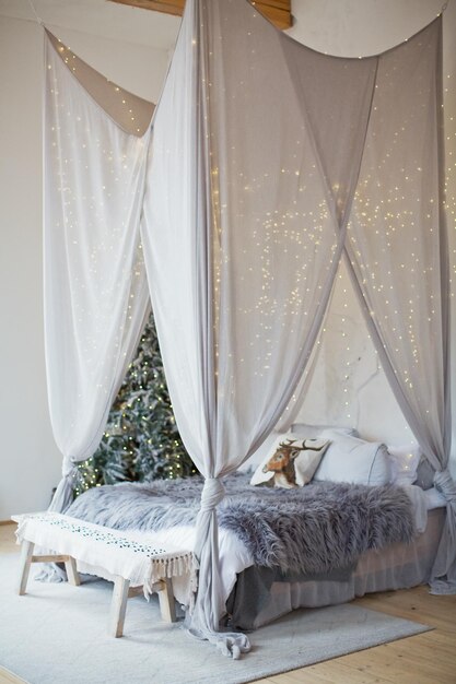 Camera da letto scandinava di Natale con baldacchino bianco con luci Albero di Natale e letto in un'accogliente stanza luminosa